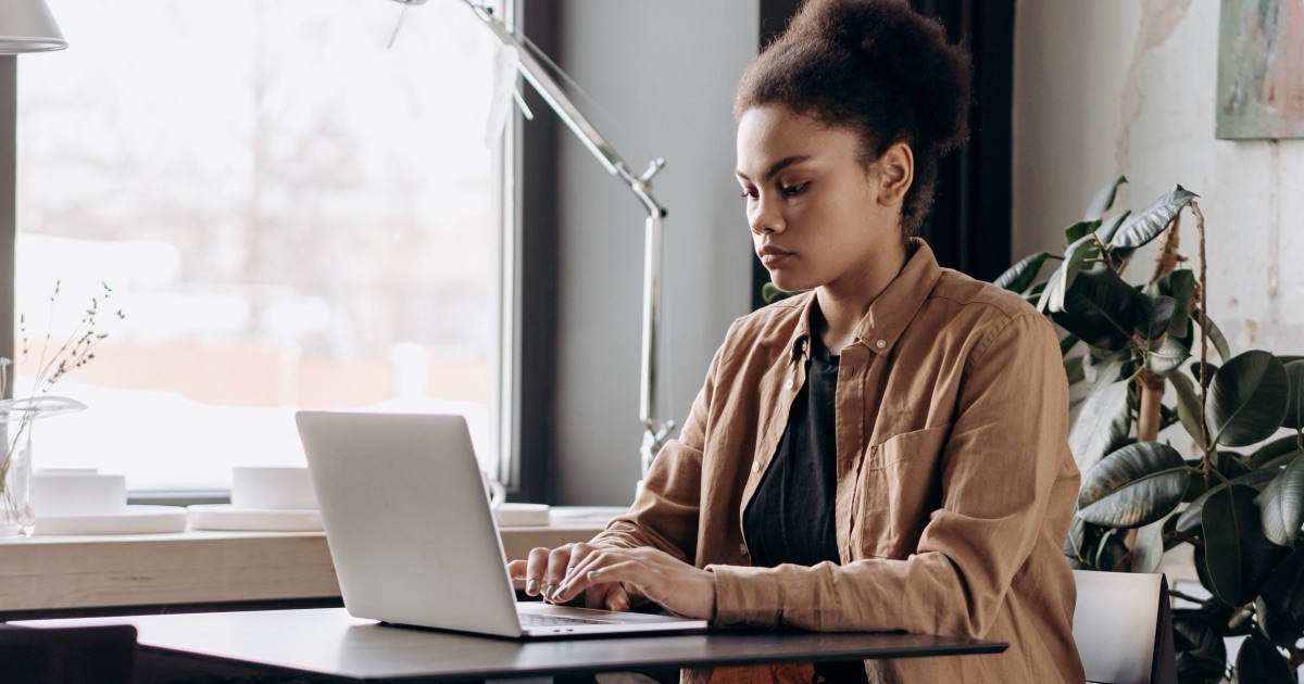 A therapist working at a laptop beside a bright office window, where the client email that Alberta PIPA and the CAP standard govern gets written and sent