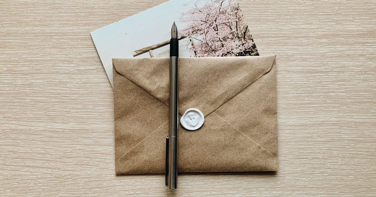 A kraft paper envelope closed with a white wax seal, a silver fountain pen laid across it and a postcard tucked behind, on a light wooden desk